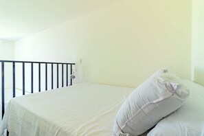 A bed with white bedding and a single white pillow, a black metal headboard, and a plain wall with a light switch. - Cupe Suites #215 - Duplex Studio by Carpediem (Ipojuca)
