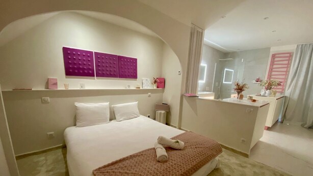 A hotel room with a large bed, a small bedside table, a mirror, and a bathroom visible through an arched doorway. - "Rose" Atypical Cocoon in the Heart of the Upper Quarter (Sète)