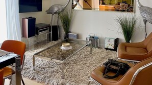 Ein modernes Wohnzimmer mit einem Glastisch, zwei Sesseln, einem Fernseher und einer durch einen großen Spiegel sichtbaren Küche.