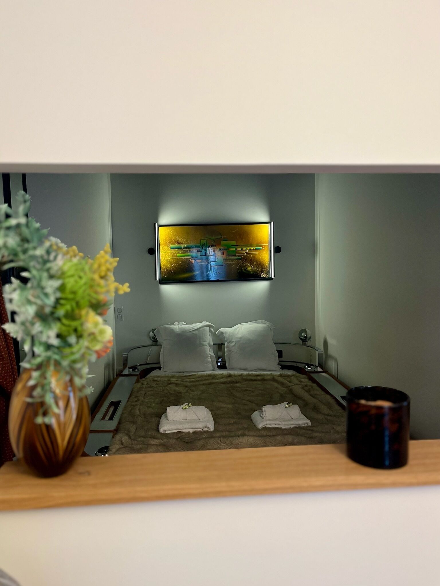 Habitació d'hotel amb un llit, dos coixins blancs, una taula de nit amb una làmpada, un vas amb flors i un televisor fixat a la paret.