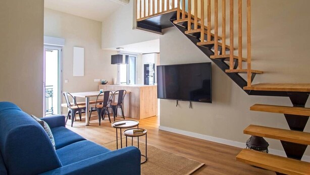 Ein modernes Wohnzimmer mit einem blauen Sofa, einem Essbereich mit Stühlen, einer Küche im Hintergrund und einer Holztreppe, an der ein Fernseher befestigt ist.