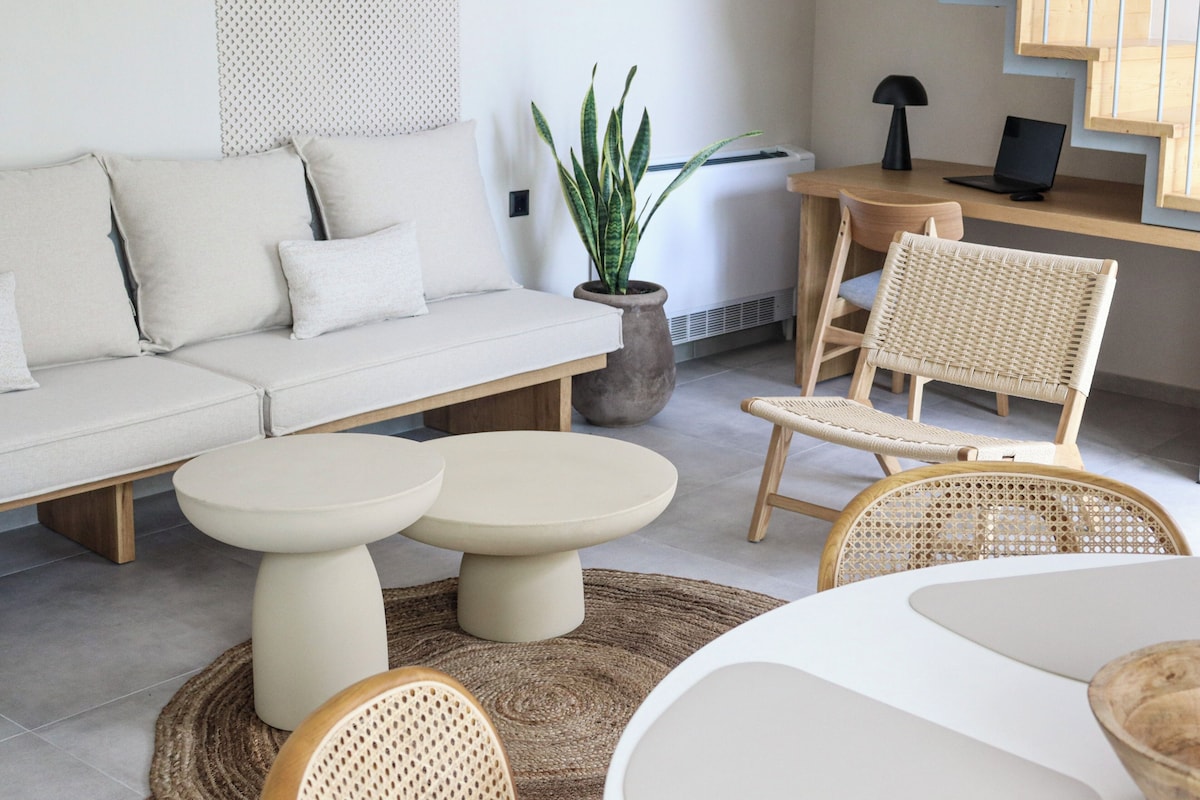 Un salon moderne avec un canapé, deux tables rondes, un fauteuil en osier, une plante en pot et un escalier en bois.