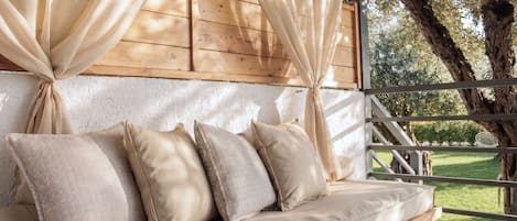 A wooden bench with cushions, a white wall, and a view of trees.