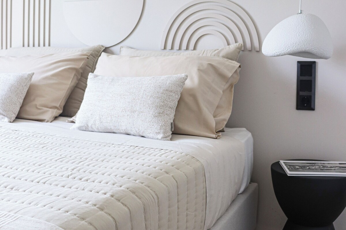A bed with a white quilted bedspread, a headboard with circular patterns, and a black side table with a lamp.