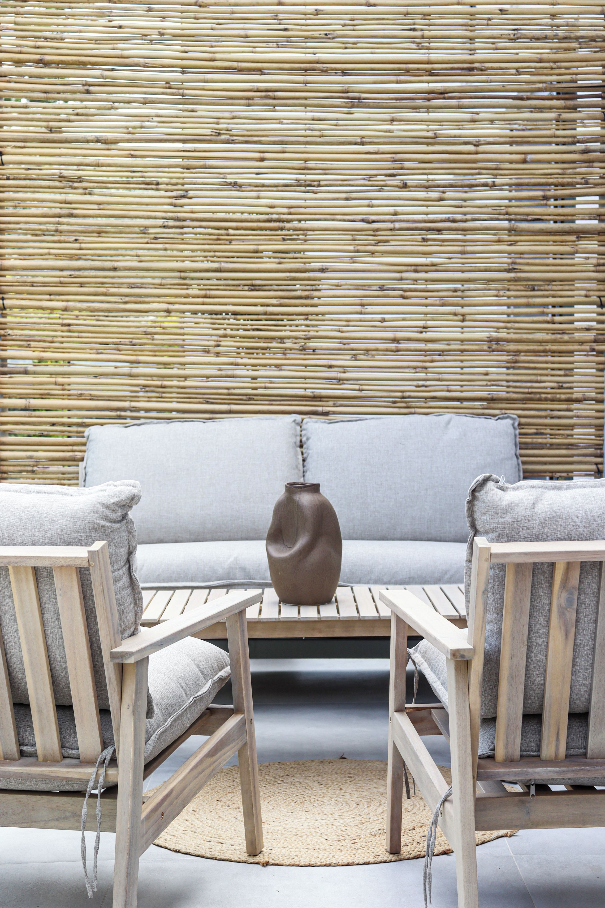 A bamboo wall, a grey sofa, two wooden chairs, a woven rug, and a vase.