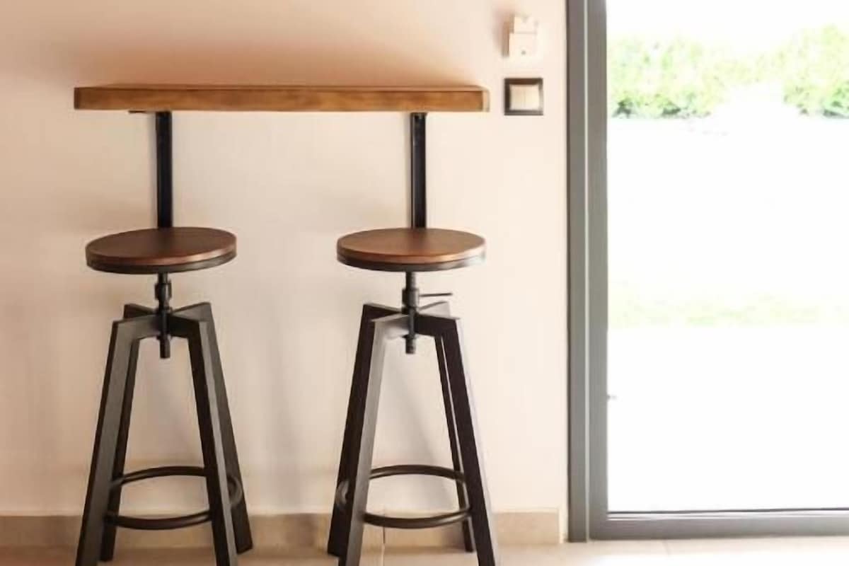 Une pièce avec un téléviseur fixé au mur, une table en bois avec deux tabourets et une porte-fenêtre coulissante donnant sur un espace extérieur.