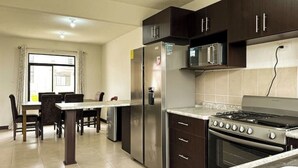 Une cuisine avec des meubles de couleur sombre, des appareils électroménagers en acier inoxydable, un coin repas avec une table et des chaises, et une fenêtre avec des rideaux.