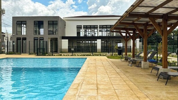 Une piscine avec une terrasse carrelée, un espace salon couvert et des bâtiments modernes en arrière-plan.