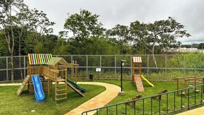 Une aire de jeux avec un toboggan et des structures d’escalade, entourée d’une clôture et de verdure.