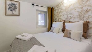 A neatly made bed with white linens and multiple pillows, a window with curtains, a framed picture of a palm tree, and a wall with a palm leaf pattern. - Neyret - Furnished apartment - Downtown Lyon (Lyon)