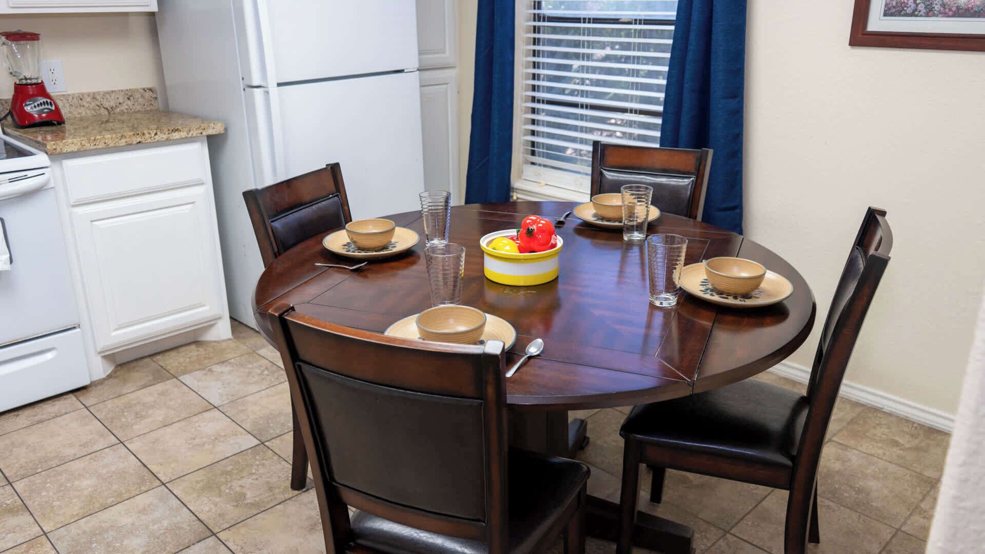 Una mesa de comedor con cuatro sillas, una licuadora roja y un refrigerador al fondo.