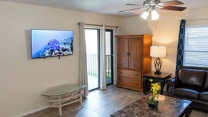 Una sala de estar con televisión, una mesa de vidrio, una mesa auxiliar con flores, un sofá de cuero, un mueble de madera, una lámpara y un ventilador de techo.