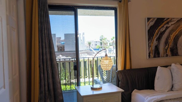 Una habitación de hotel con una cama, una mesita de noche, una lámpara y vistas de la ciudad a través de un ventanal grande.