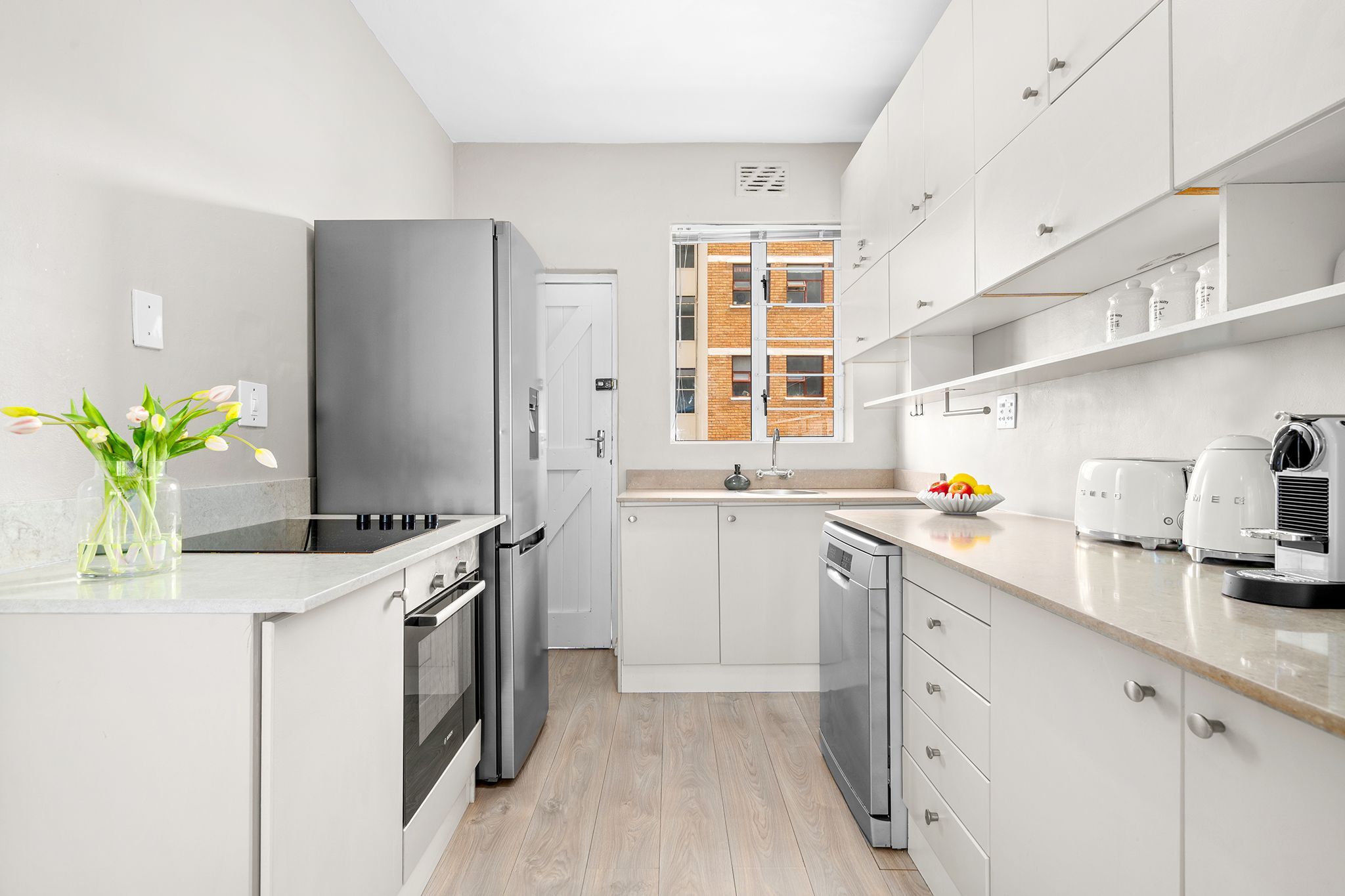 Una cocina moderna con armarios blancos, electrodomésticos de acero inoxidable y vista a un edificio de ladrillo a través de una ventana.