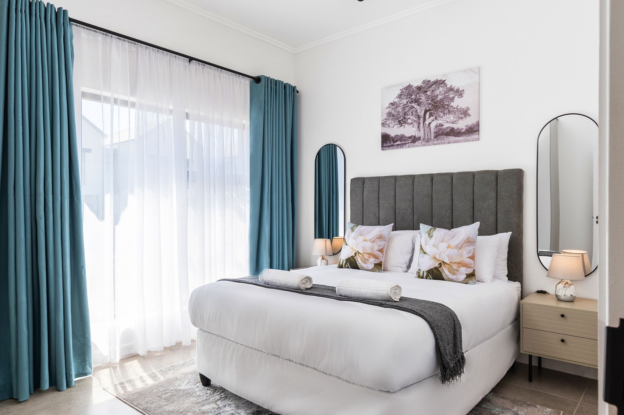 A bedroom with a bed, a bedside table, a lamp, a mirror, and a window with curtains.