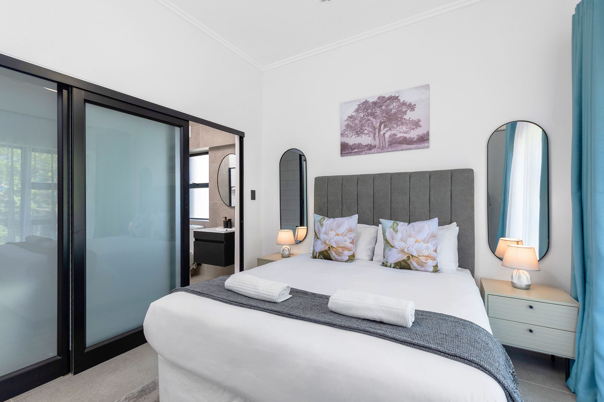 A modern hotel room with a large bed, a sliding glass door, a nightstand with a lamp, and a painting on the wall.