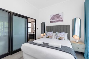 A modern hotel room with a large bed, a sliding glass door, a nightstand with a lamp, and a painting on the wall.
