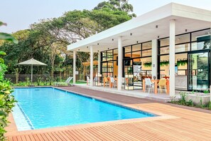 A modern pool area with a wooden deck, outdoor seating, and a covered patio with glass walls. - Woodlands Estate 22 (Ballito)