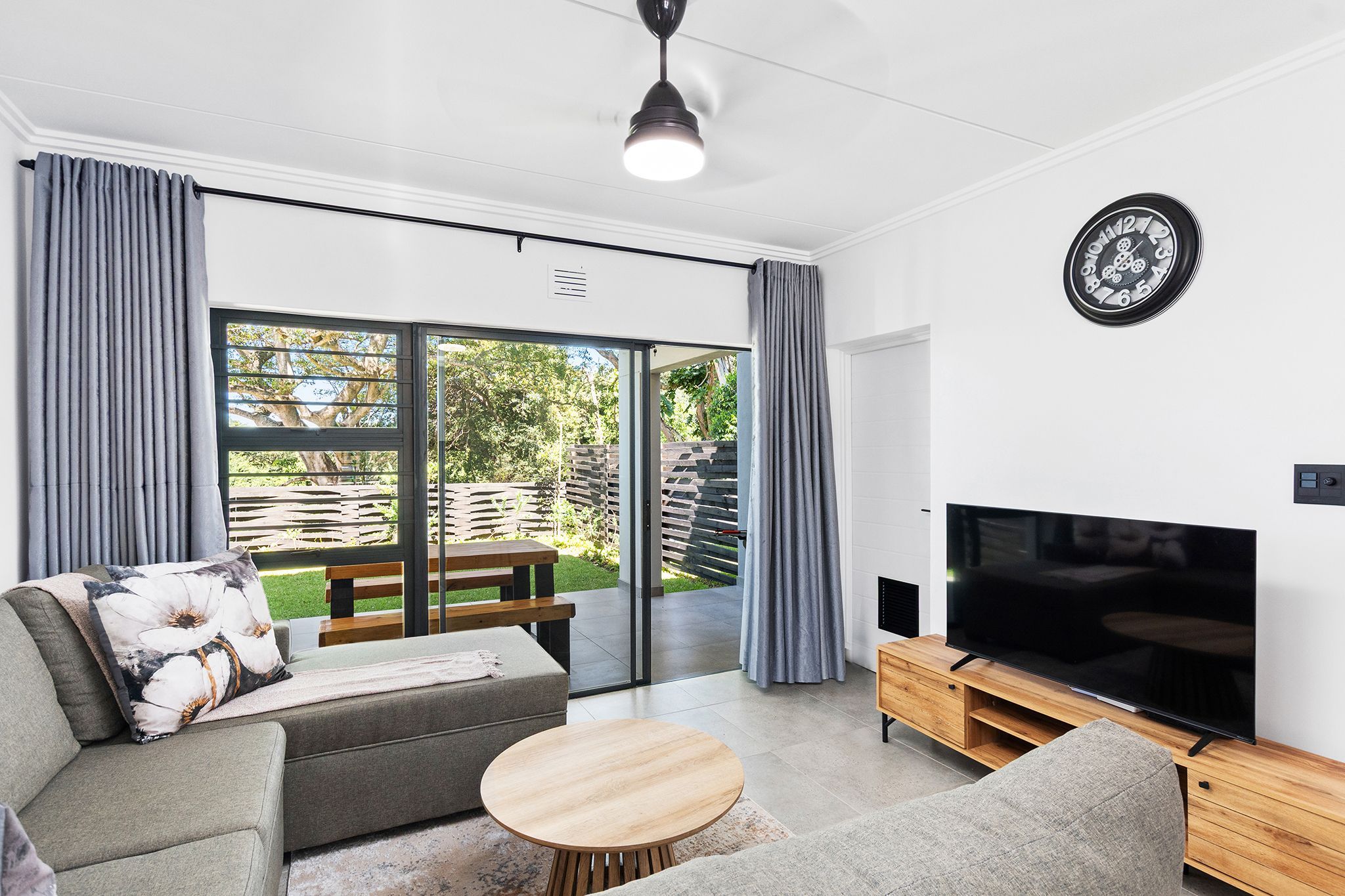 A modern living room with a sofa, a coffee table, a TV on a wooden stand, and a sliding glass door leading to an outdoor area.