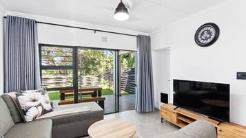 A modern living room with a sofa, a coffee table, a TV on a wooden stand, and a sliding glass door leading to an outdoor area.