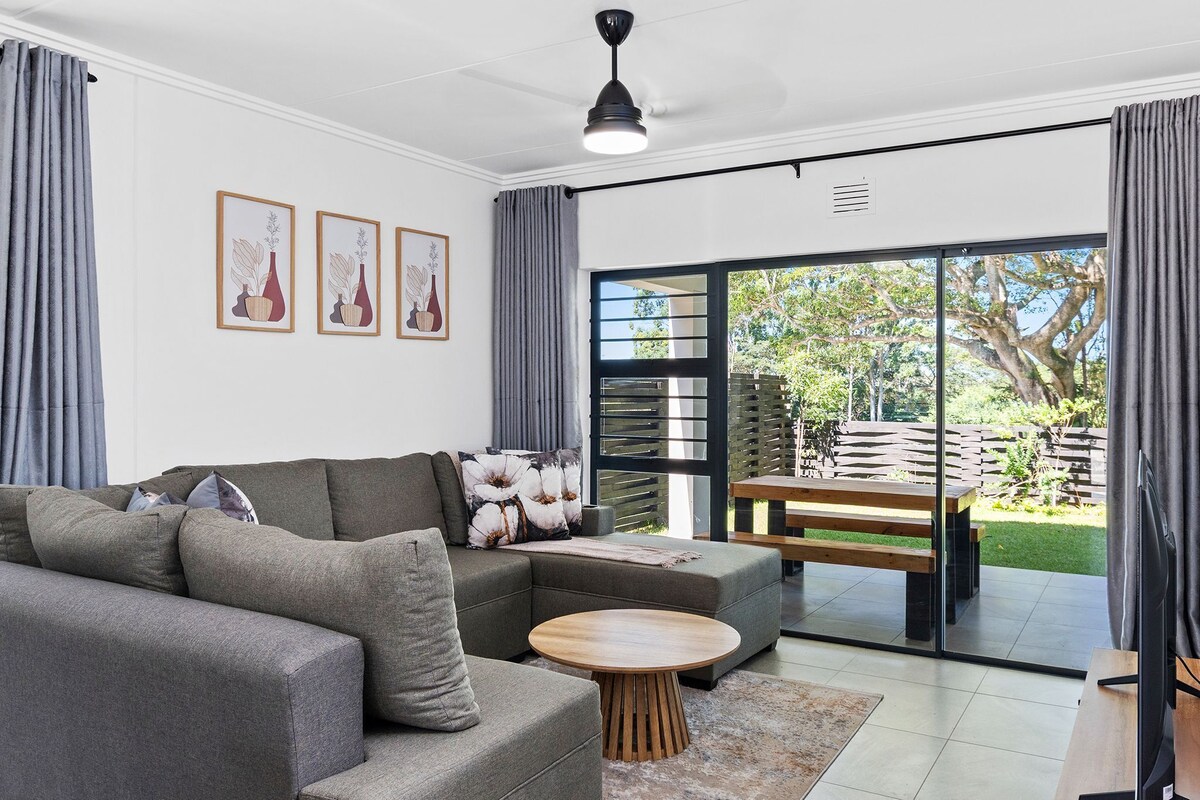 A modern living room with a sectional sofa, a round wooden coffee table, and a sliding glass door leading to a garden.