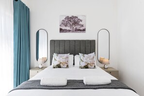 A neatly made bed with pillows, a bedside table with a lamp, a wall-mounted mirror, and a picture of a tree.