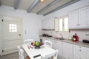Une cuisine avec des meubles blancs, une table à manger avec des chaises, un panier de fruits, une bouilloire et une machine à café.
