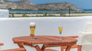Ein Holztisch mit zwei Gläsern Getränke, ein weißer Stuhl und im Hintergrund Meer und Berge.