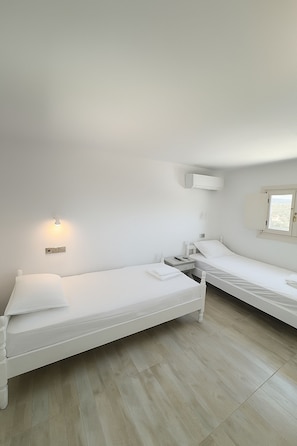 A small, clean room with two single beds, a bedside table with a lamp, a window, and a wall-mounted air conditioner. - My Villa Naxos I (Naxos)