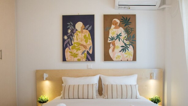 A neatly made bed with two rolled towels, a wooden headboard, two wall-mounted night lights, and two framed floral paintings above the bed. - K11 Apartment (Athens)