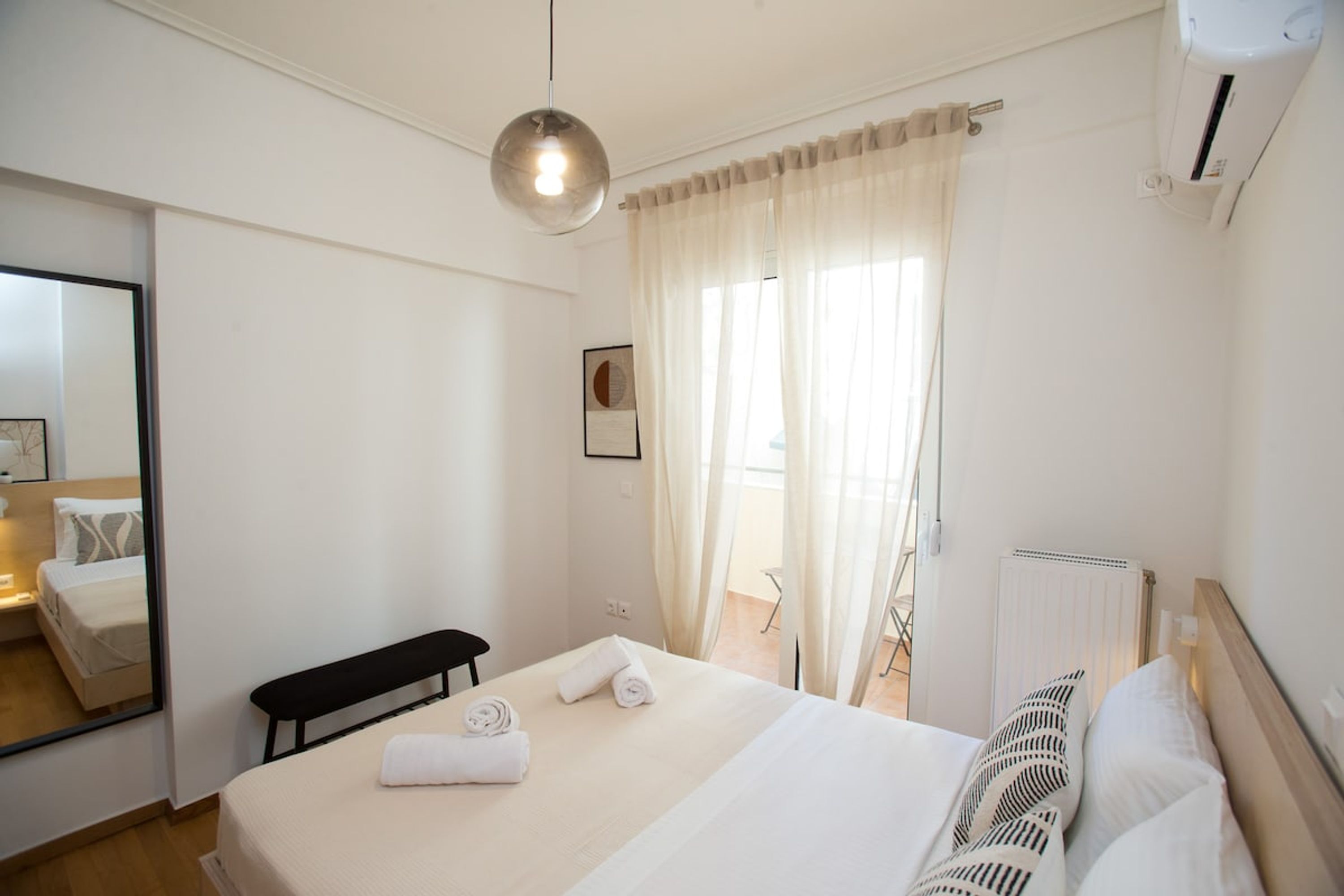 A hotel room with a bed, a bench, a mirror, a window with curtains, and an air conditioning unit.