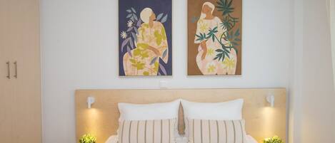 A bedroom with a bed, two nightstands, two wall-mounted lamps, and two wall art pieces.
