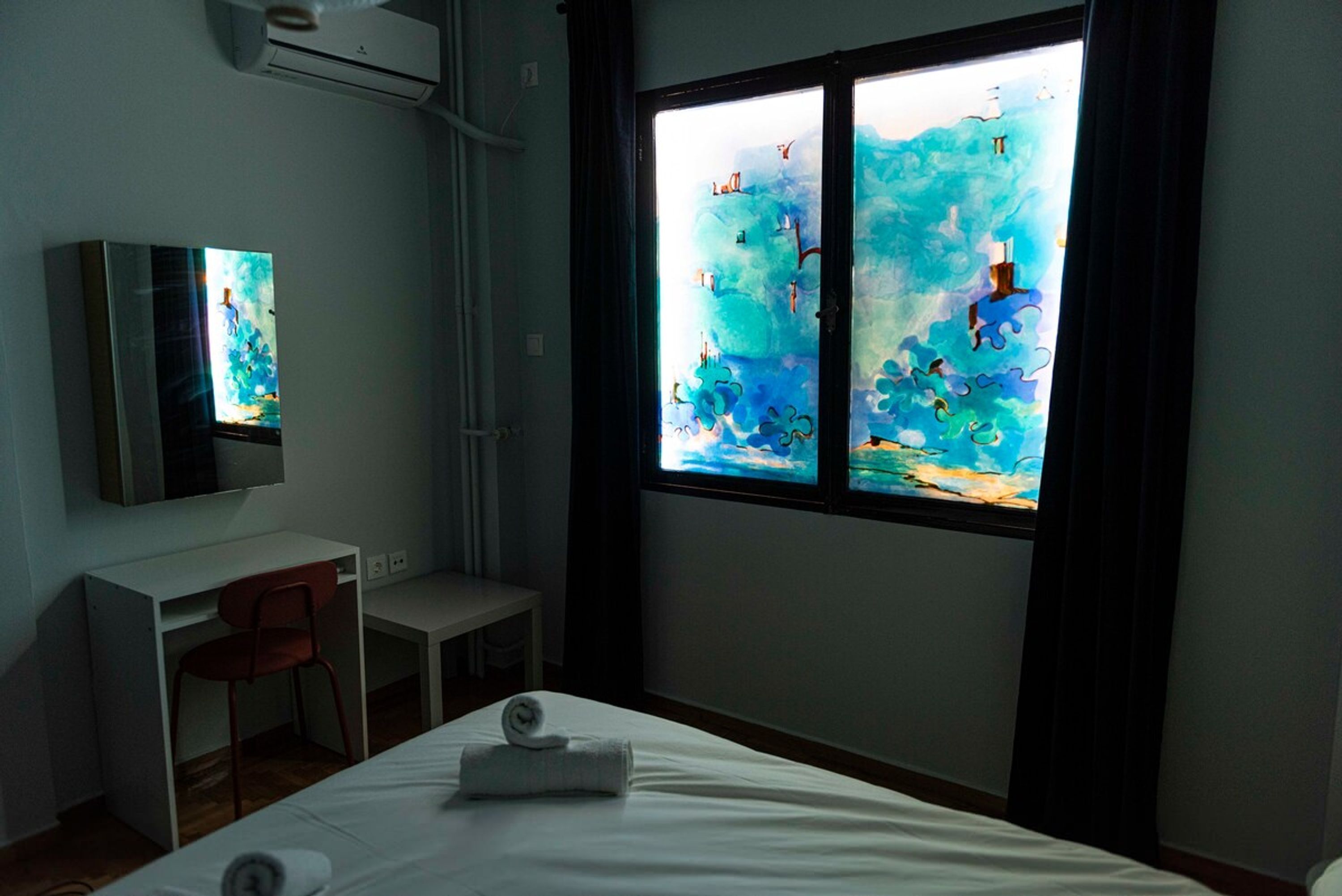 A hotel room with a bed, a desk, a chair, and a large window with a view of the sky and clouds.
