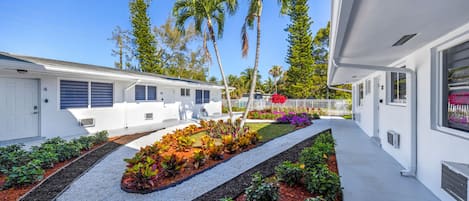 A modern building with a white exterior, a well-maintained garden with various plants and a gravel pathway, and tall palm trees in the background.