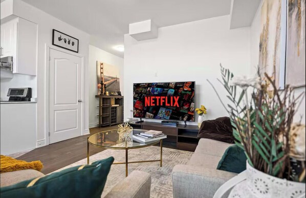 A modern living room with a flat-screen TV displaying 'NETFLIX', a glass coffee table, and a sofa. - Cozy Comfort Escape - Your Next Memorable Retreat (Brampton)