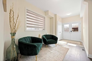 A modern living room with two dark green armchairs, a large window with blinds, a white door, and a decorative vase with dried plants. - Cozy Comfort Escape - Your Next Memorable Retreat (Brampton)