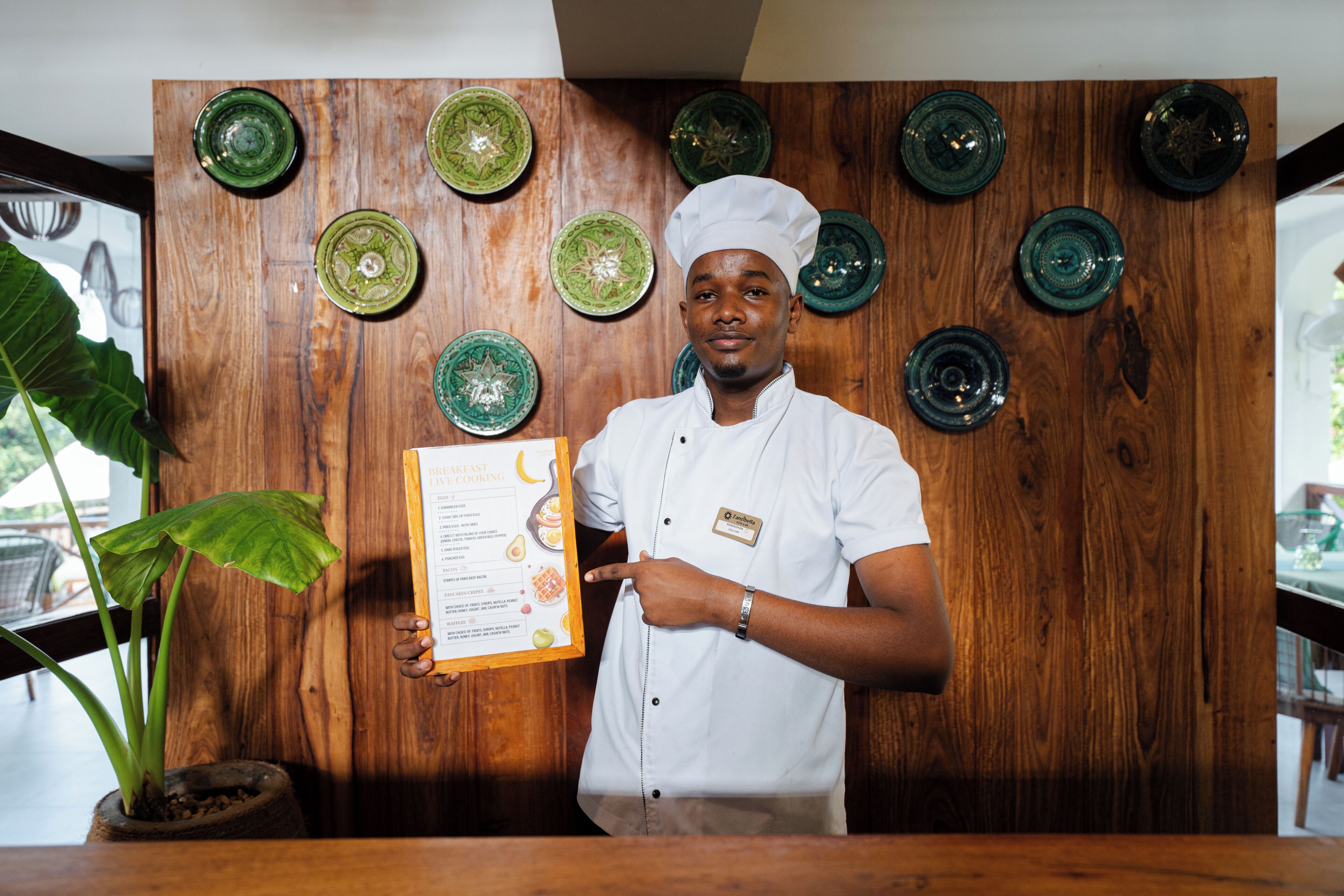Una persona con uniforme blanco de chef sostiene un menú enmarcado, frente a una pared de madera con platos decorativos verdes.