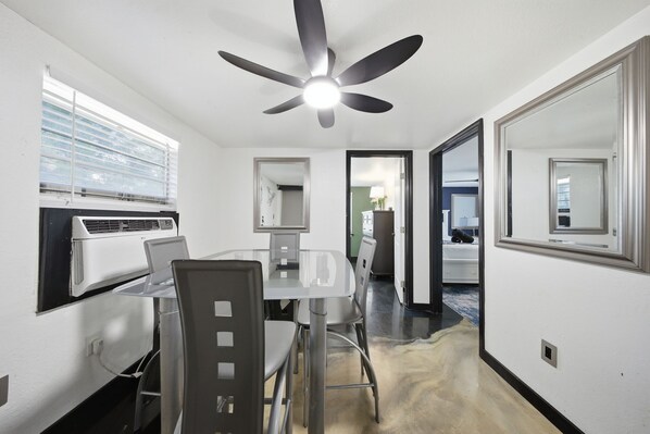 A compact hotel room with a dining table, chairs, a ceiling fan, a window with blinds, and a mirror on the wall.