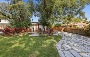 Una propiedad residencial con entrada pavimentada, un pequeño jardín delantero con plantas rojas y una valla de madera.