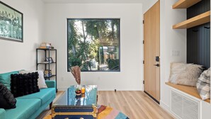 Ein modernes Wohnzimmer mit einem petrolfarbenen Sofa, einem Glastisch und einem großen Fenster mit Blick auf Bäume.