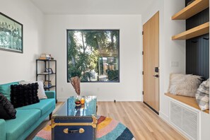 Ein modernes Wohnzimmer mit einem petrolfarbenen Sofa, einem Glastisch und einem großen Fenster mit Blick auf Bäume.