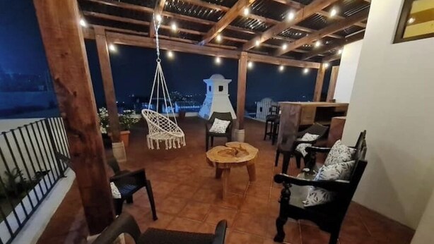 Un patio exterior con muebles de madera, una silla colgante y vistas de la ciudad de noche.
