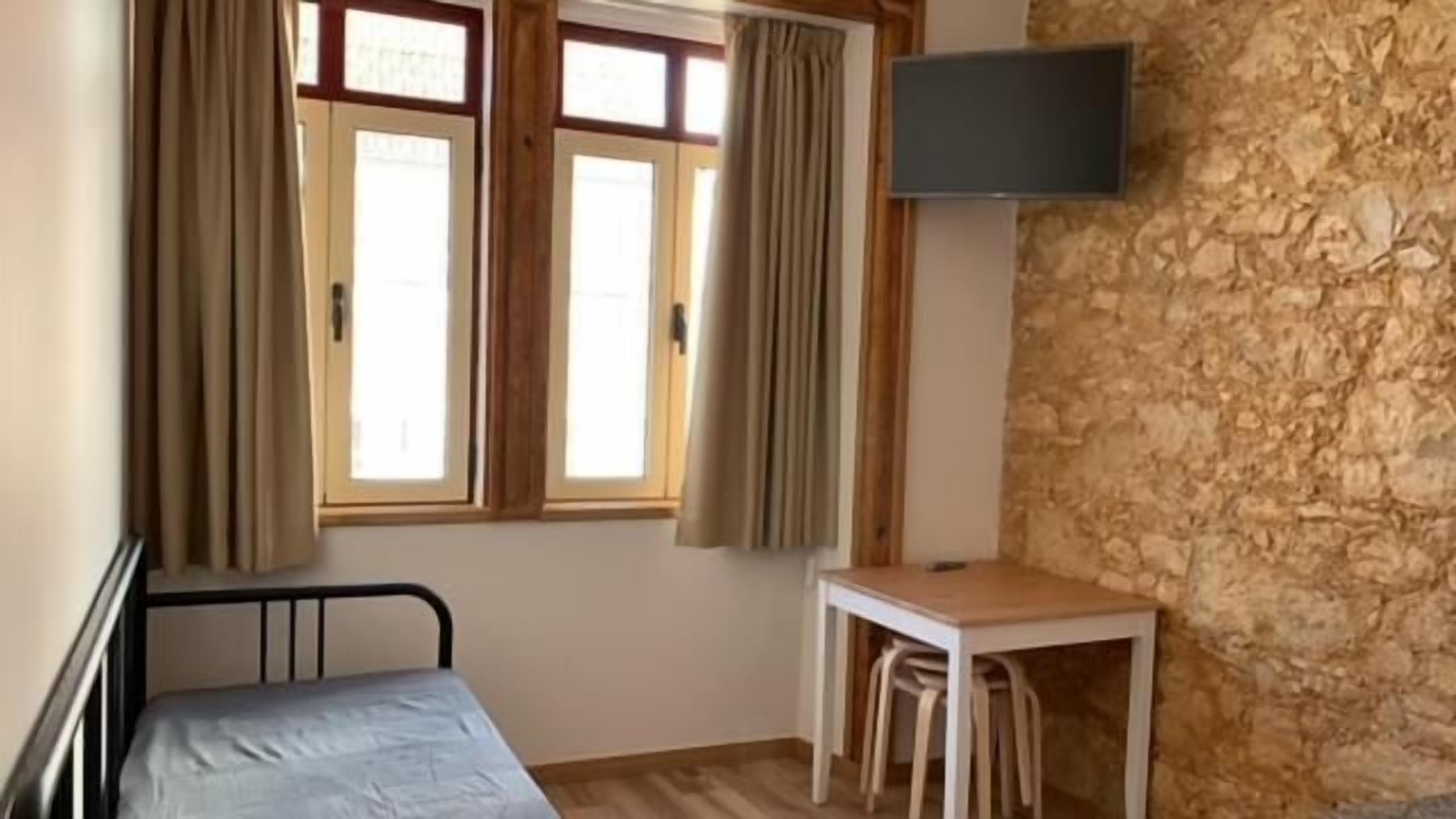 Une pièce comprenant un lit, une table avec des chaises, un téléviseur fixé au mur et une fenêtre avec des rideaux.