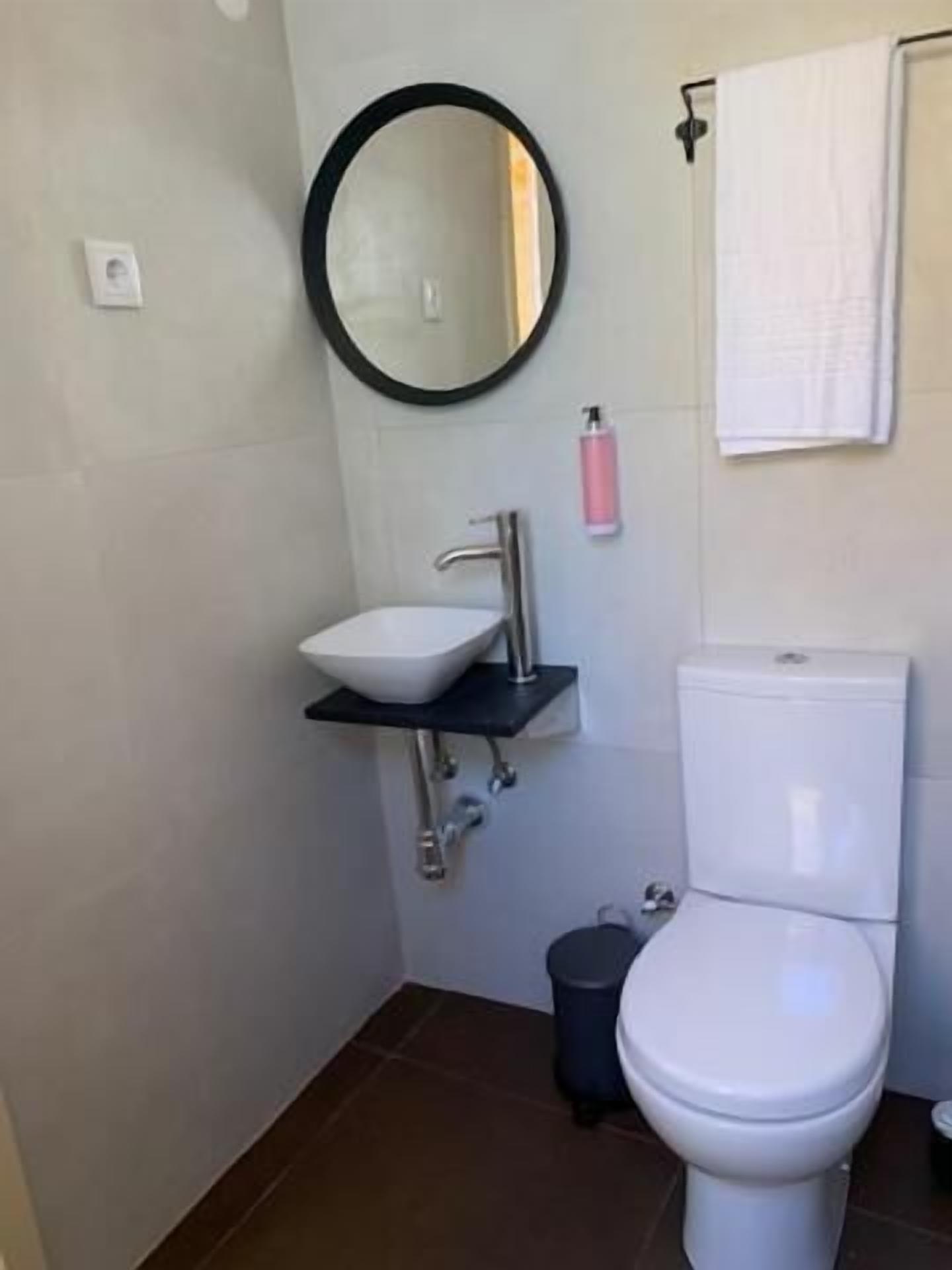 Une salle de bain avec un lavabo blanc, un miroir rond, des toilettes et une poubelle.