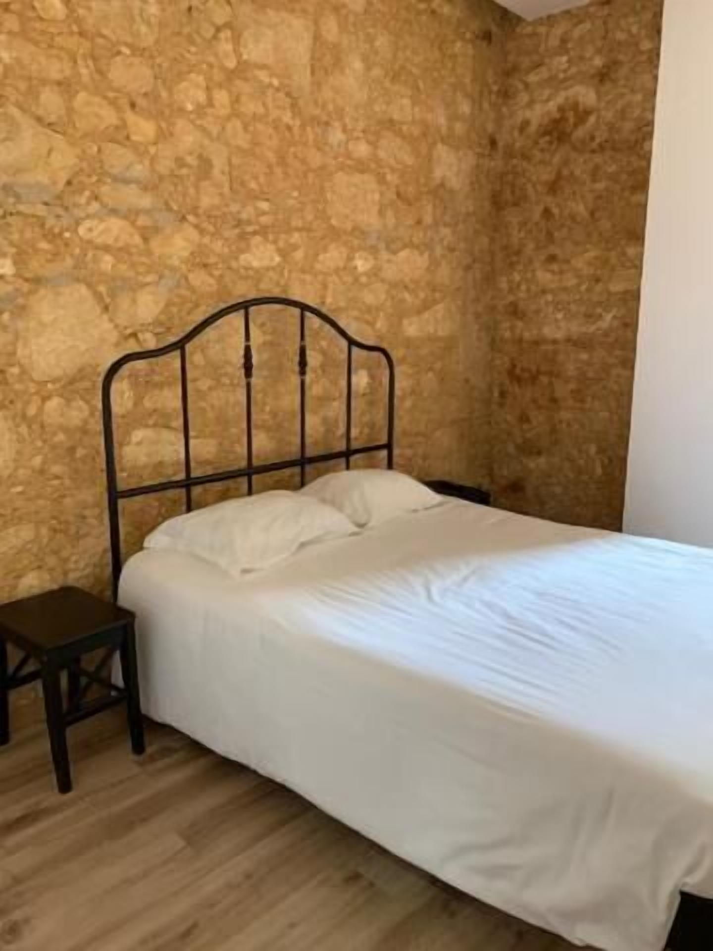 Een bed met metalen frame, witte beddengoed en een klein houten nachtkastje ernaast. De kamer heeft een stenen muur en houten vloer.