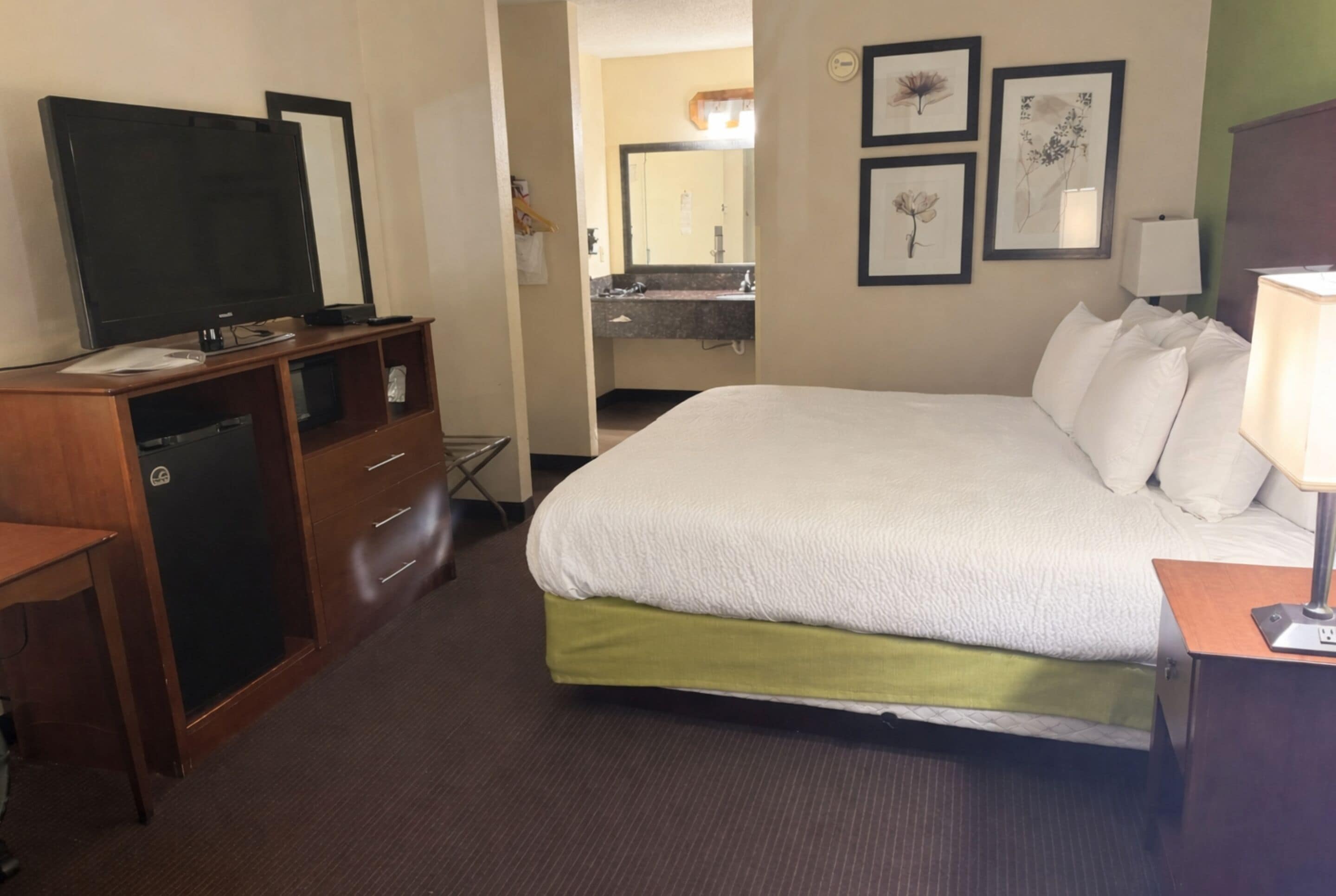 A hotel room with a bed, a desk, a television, a bathroom with a sink and mirror, and framed pictures on the wall.
