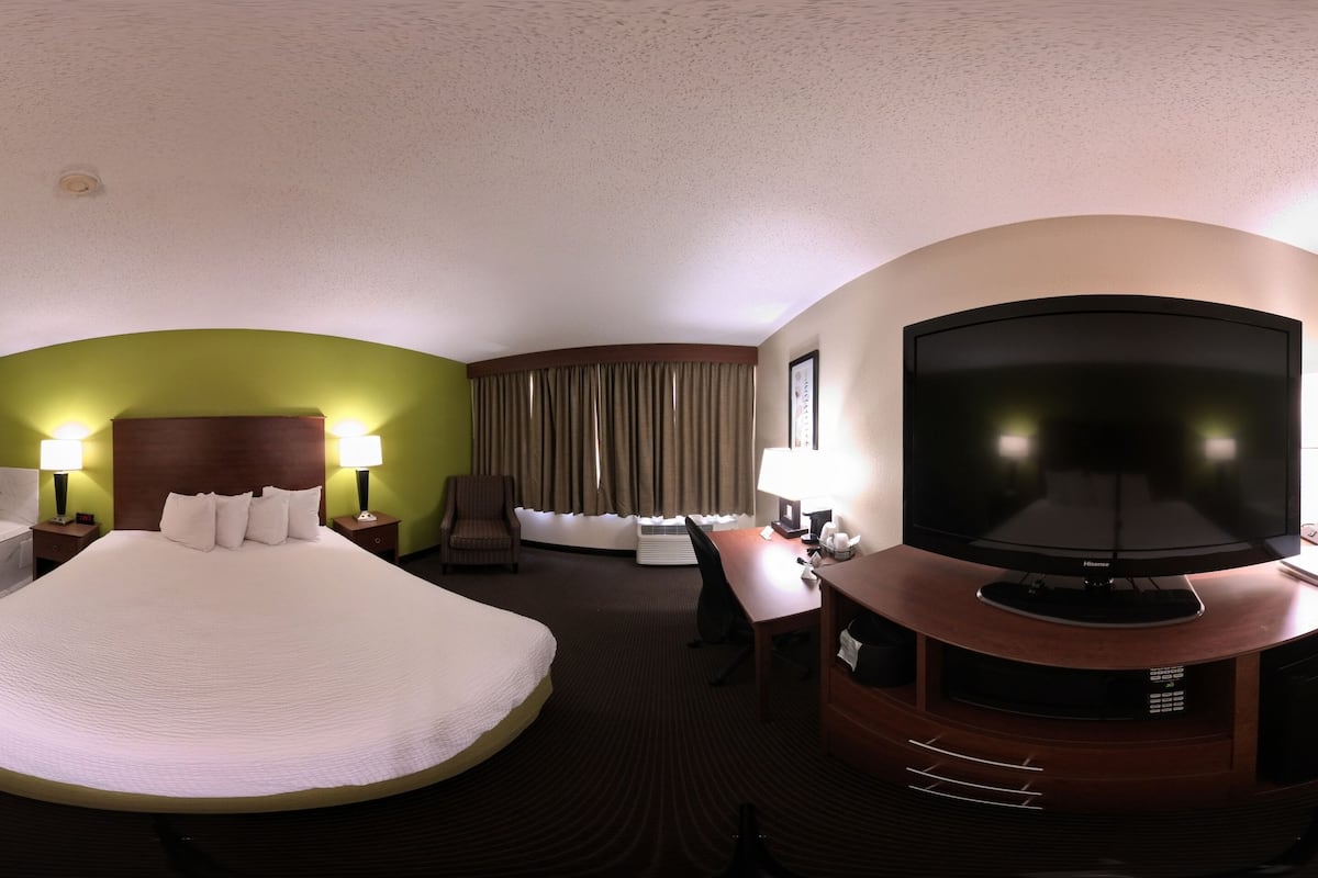 A hotel room with a large bed, a desk with a flat-screen TV, a chair, and a desk lamp.