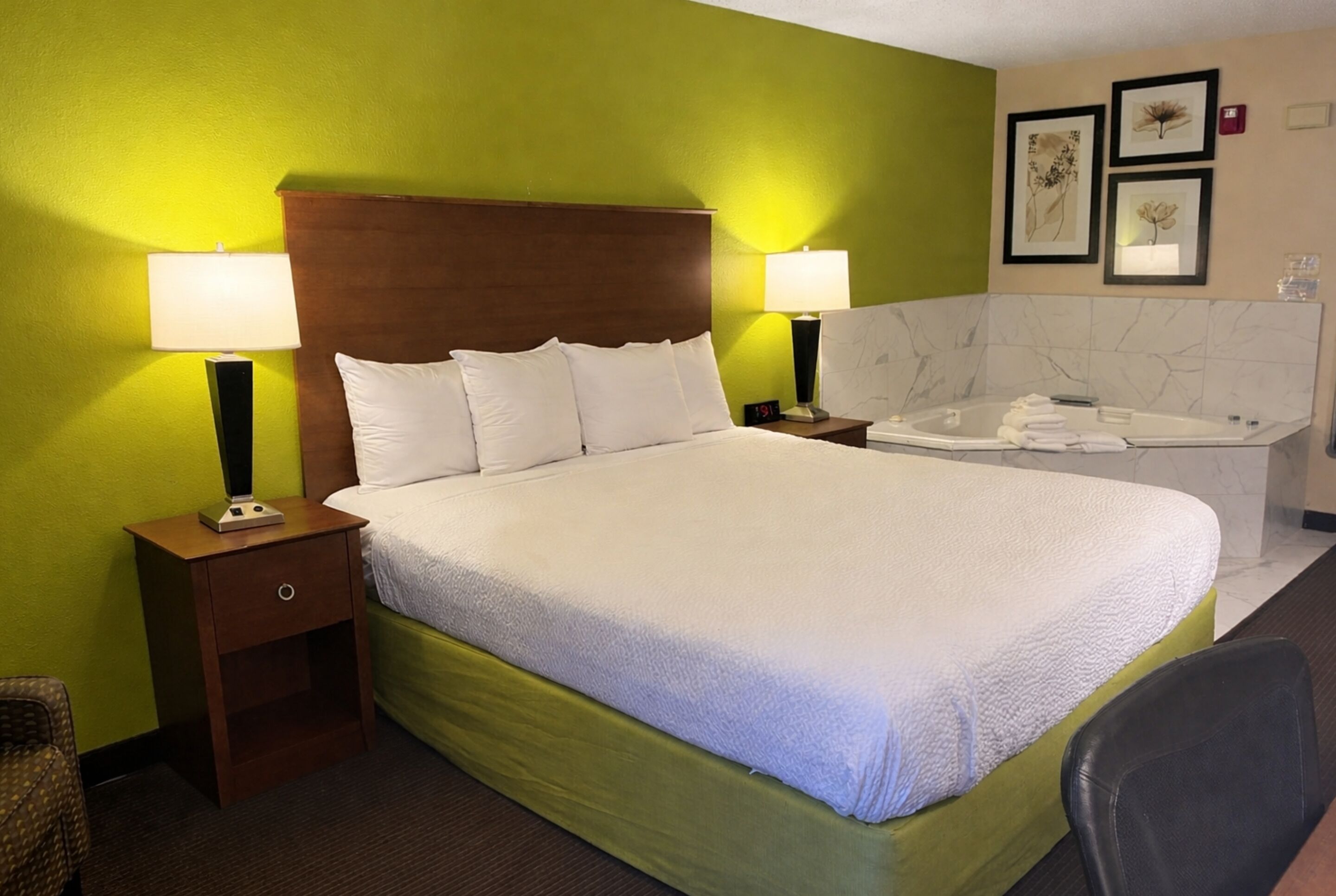 A hotel room with a bed, two bedside lamps, a nightstand, a chair, a bathroom with a bathtub, and a marble wall.