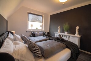 A bedroom with two beds, a window with blinds, a bedside table with a lamp, and decorative items on a shelf. - Cozy apartment in Emden, 5 minutes to the city center (Emden)