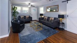 A living room with a dark grey sofa, a black armchair, a glass coffee table, and a blue striped rug. There are two ceiling fans, a large mirror, and a sliding barn-style door. - Charming 3BR Retreat in Plaza Close to Uptown (Charlotte)